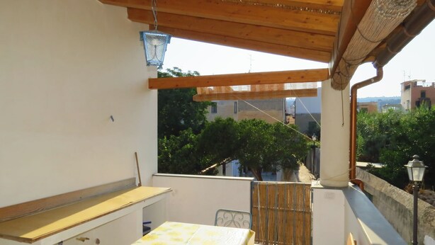 Un balcón con una mesa y sillas de madera, un farol azul colgante, y vistas a zonas verdes y edificios al fondo.