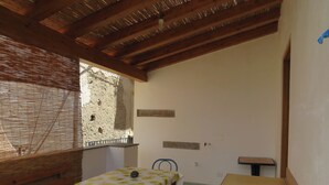 Una habitación con techo de madera, una mesa con mantel floral, un banco y una ventana pequeña con vista a un muro de piedra.