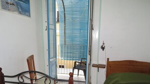 A room with a bed, a chair, a table, and a door leading to an outdoor area with a blue railing. - Deluxe Double (Lipari)