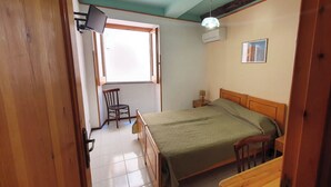 A bedroom with a wooden bed, a chair, a bedside table, a window with a view, and an air conditioning unit. - double use room on the street floor (Lipari)