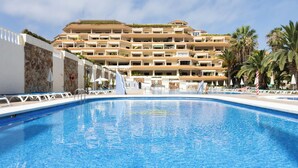 Une piscine entourée d’une terrasse carrelée, elle-même bordée d’un hôtel à plusieurs étages doté de balcons et de palmiers.