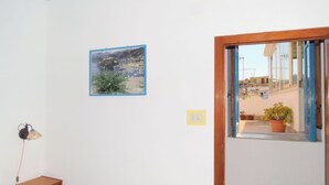 Une chambre à coucher comprenant un lit, une table de chevet, une lampe fixée au mur, une petite plante en pot et une fenêtre donnant sur des immeubles et une rue.