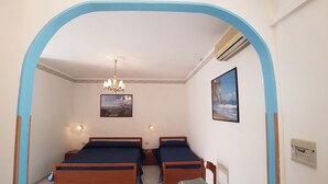 A bedroom with two wooden beds, a chandelier, a framed picture of a beach, and an air conditioning unit.