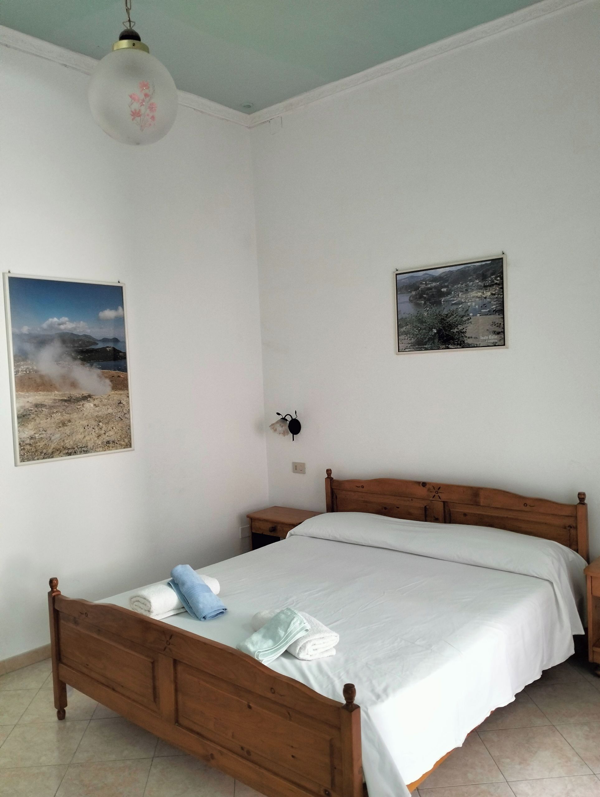 A bedroom with a wooden bed, white bedding, a wall-mounted light fixture, and two framed pictures.