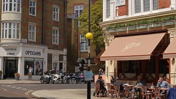 Une scène de rue avec un café appelé « Sautine » et un bâtiment portant l’enseigne « SIMON OPTICIENS ».