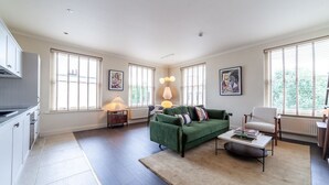 Un vaste salon avec un canapé vert, une table basse blanche et un fauteuil blanc. De grandes fenêtres sont équipées de stores, et on y trouve également un radiateur ainsi qu’un coin cuisine avec des meubles blancs.