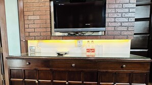 A wooden cabinet with drawers, a flat-screen TV mounted on a brick wall, and a shelf with bottled water. - Qotel Signature Lavish Inn Rajouri Garden (New Delhi)
