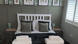 A bed with white headboard, grey and white pillows, and a black bedspread. There are framed botanical prints on the wall above the bed. - 1-bed Apartment - Sleeps 2 & Private Parking (Llansantffraid-ym-Mechain)