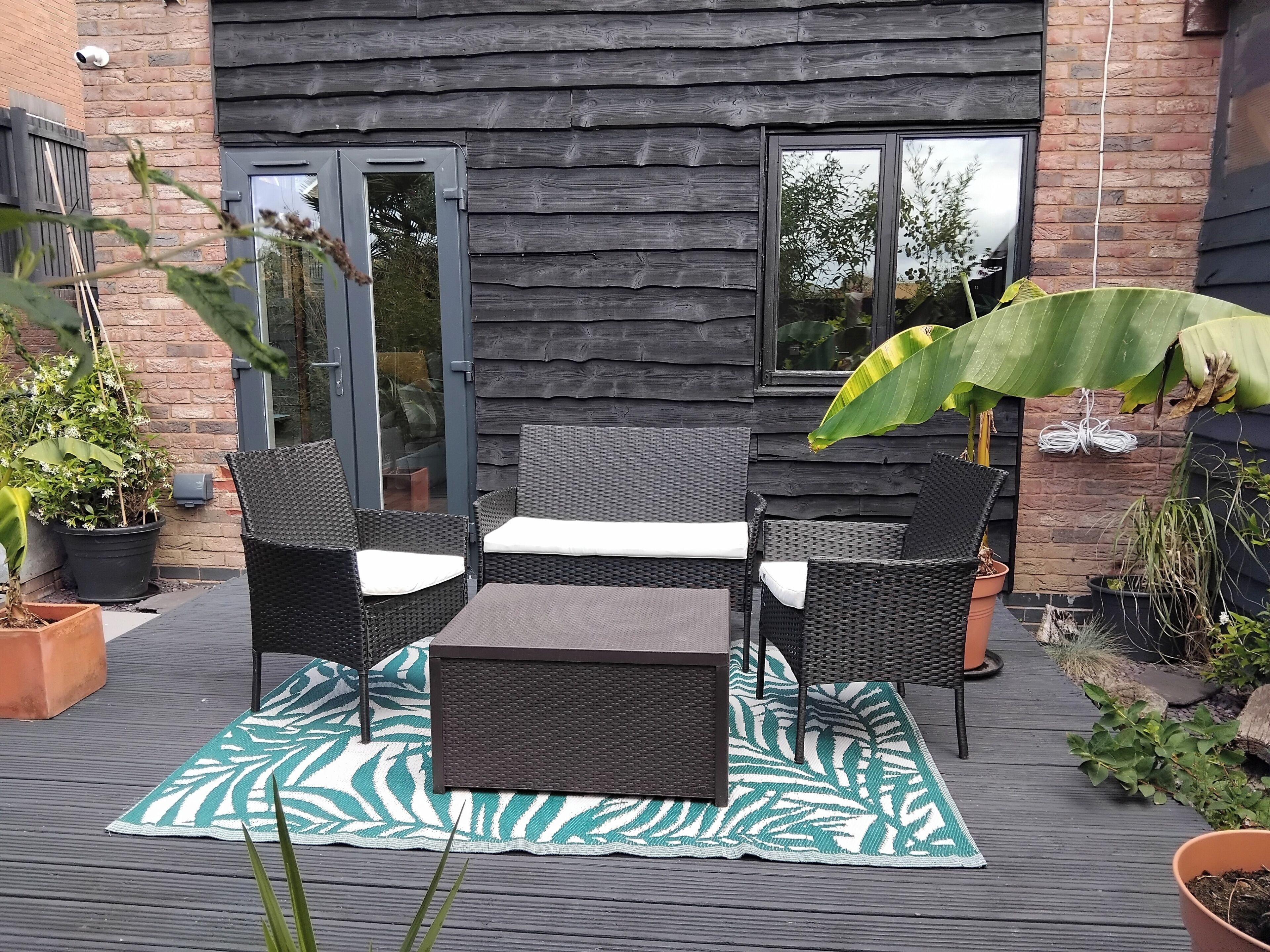 An outdoor patio with wicker furniture, a green and white patterned rug, and potted plants.