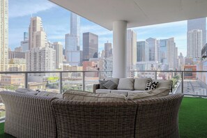 A balcony with a wicker sofa and cushions, overlooking a cityscape with tall buildings.