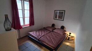 A bedroom with a bed, a window with curtains, a framed picture on the wall, and a bedside lamp.