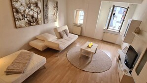 A modern living room with a sofa, a small table, and a TV. There are two windows providing natural light.