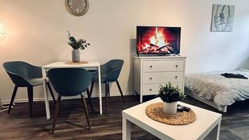 A cozy room with a TV displaying a fireplace, a dining table with chairs, a small white table with a plant, and a bed with a blanket.
