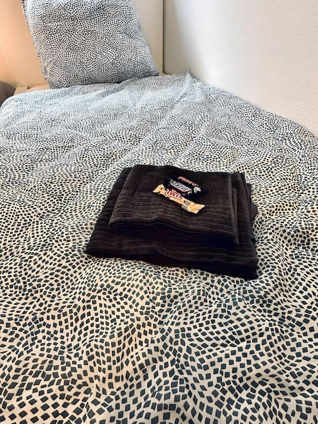 Een netjes opgevouwen zwart T-shirt met zichtbaar logo op een bed met een geborduurd sprei.