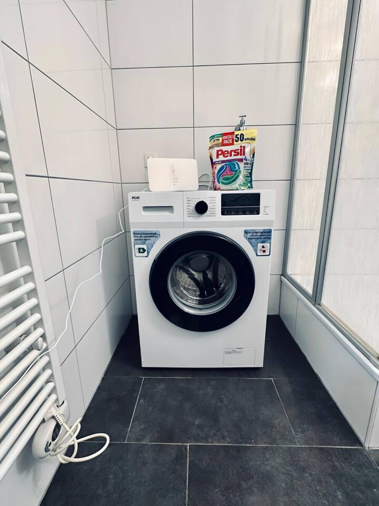A compact, modern laundry room with a front-loading washing machine, a drying rack, and a bag of Persil detergent.