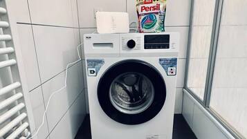 A compact, modern laundry room with a front-loading washing machine, a drying rack, and a bag of Persil detergent.