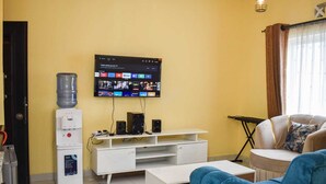 Una sala de estar con un televisor de pantalla plana montado en la pared, un dispensador de agua, un mueble blanco para entretenimiento, un sofá azul y un sillón beige.