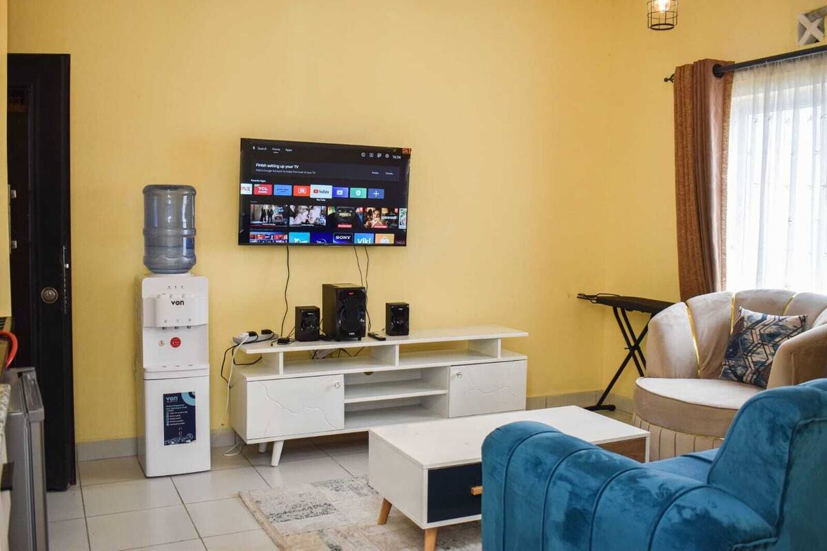 Una sala de estar con un televisor de pantalla plana montado en la pared, un dispensador de agua, un mueble blanco para entretenimiento, un sofá azul y un sillón beige.