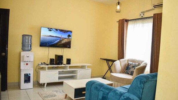 Una sala de estar con un televisor de pantalla plana montado en la pared, un mueble blanco para equipo de sonido con altavoces, un sofá azul, una mesa de centro blanca y un dispensador de agua.