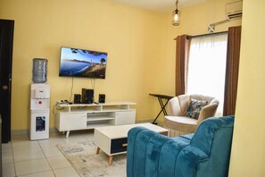 A living room with a flat-screen TV mounted on the wall, a white entertainment unit with speakers, a blue sofa, a white coffee table, and a water dispenser. - Lux Suites Buxton Point Apartments (Mombasa)