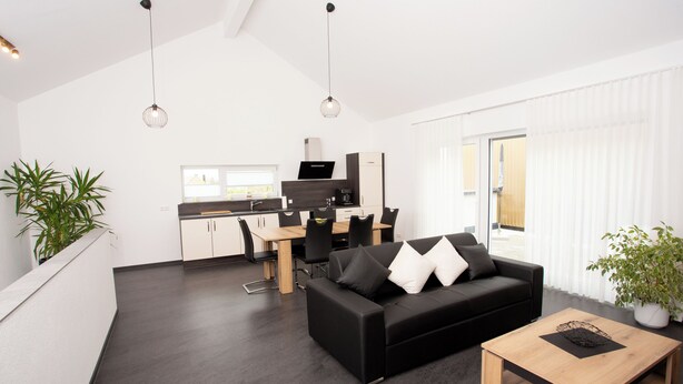 Ein modernes Wohnzimmer mit einem schwarzen Sofa, einem Holztisch und einem Essbereich, im Hintergrund befindet sich eine Küche.