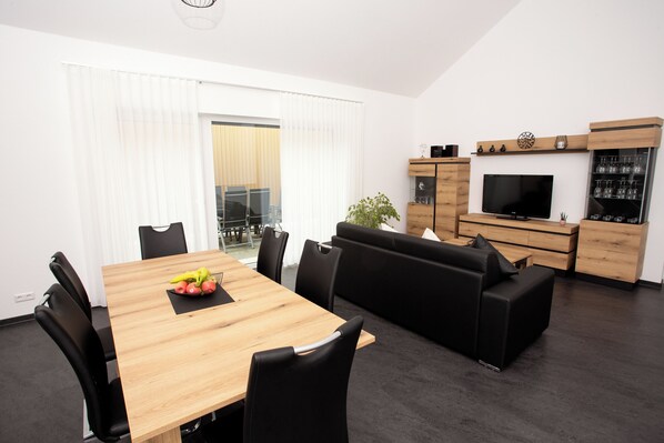 A modern living room with a wooden dining table, black chairs, a black sofa, a TV, and a wine cabinet.