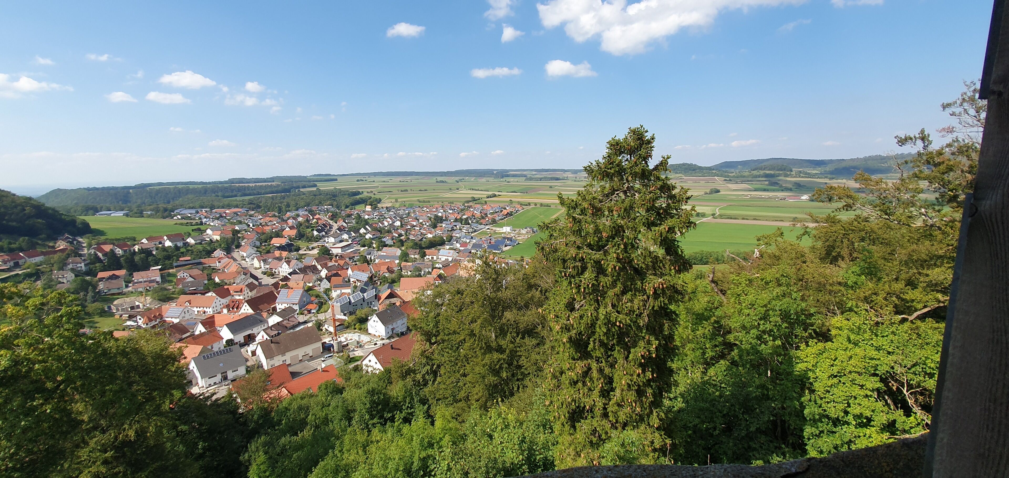 A panoramic view of a town with red-roofed houses, surrounded by greenery and hills.