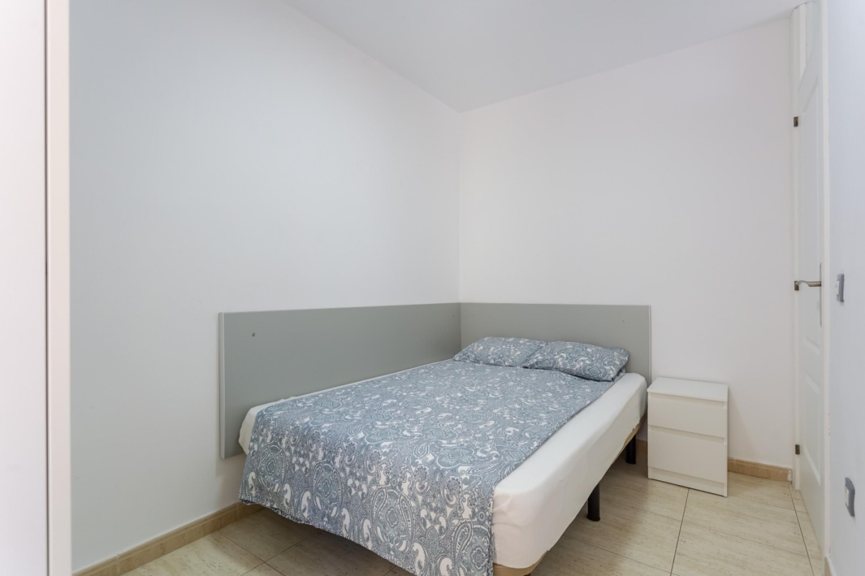 A small, single-bed room with a grey headboard, a bedside table, and a white drawer unit.