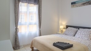 Un dormitorio con una cama, una mesita de noche con lámpara, una ventana con cortinas y un cuadro en la pared.