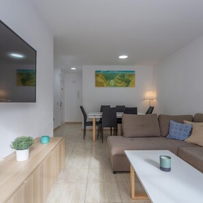 Ein modernes Wohnzimmer mit einer Couch, einem Couchtisch, einem an der Wand montierten Fernseher, einem Essbereich mit Stühlen und einem Tisch sowie einem Bild an der Wand.