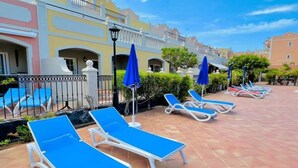 A poolside area with sun loungers and umbrellas, a tiled floor, and a building with balconies in the background. - Enjoy 1 BR Palm Mar Next to Pool (Arona)
