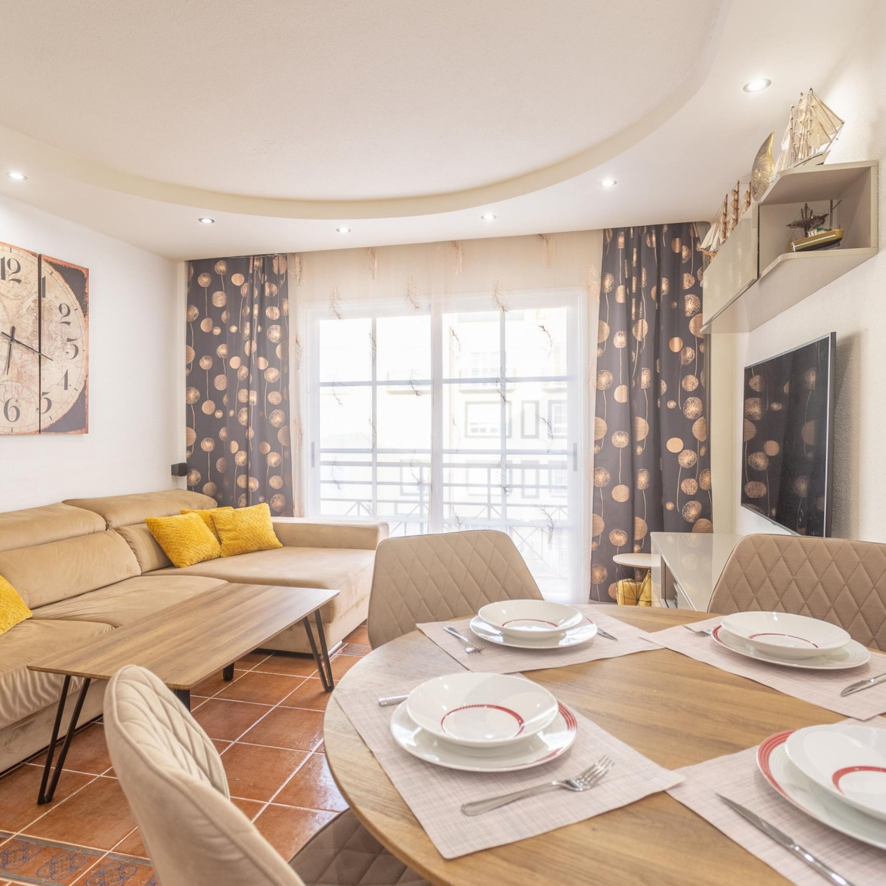A dining area with a round table set for four, a sofa with yellow cushions, and a large wall clock.