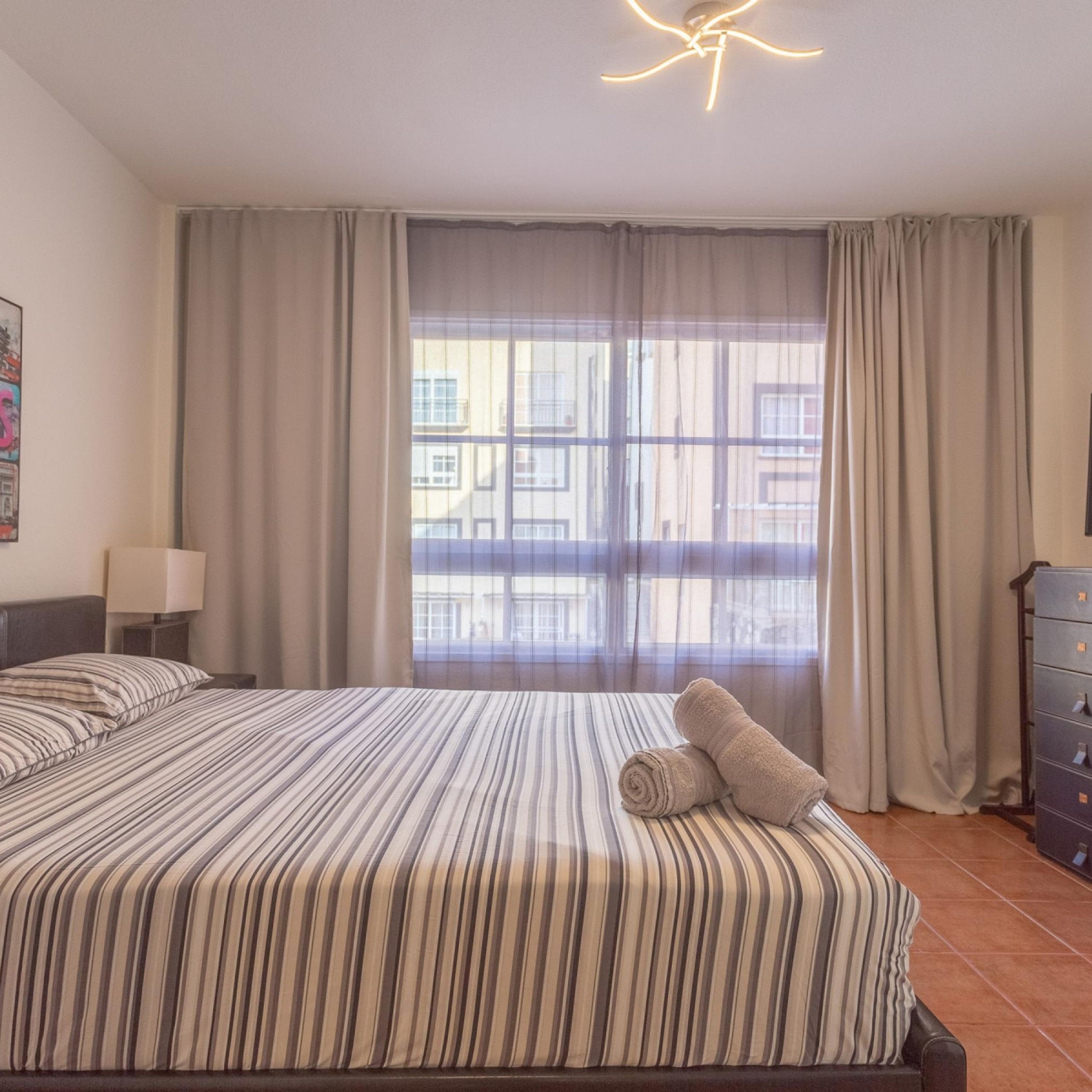 A hotel room with a large bed, a nightstand, a lamp, a dresser, and a view of buildings through sheer curtains.
