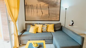 A living room with a gray sofa, yellow cushions, a yellow table with a bowl of fruit, and a large beach-themed painting on the wall.