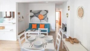 A modern dining area with a white table set for four, a blue sofa with orange cushions, and a stone wall art piece. - Central 2bedroom 200m From Beach (Adeje)