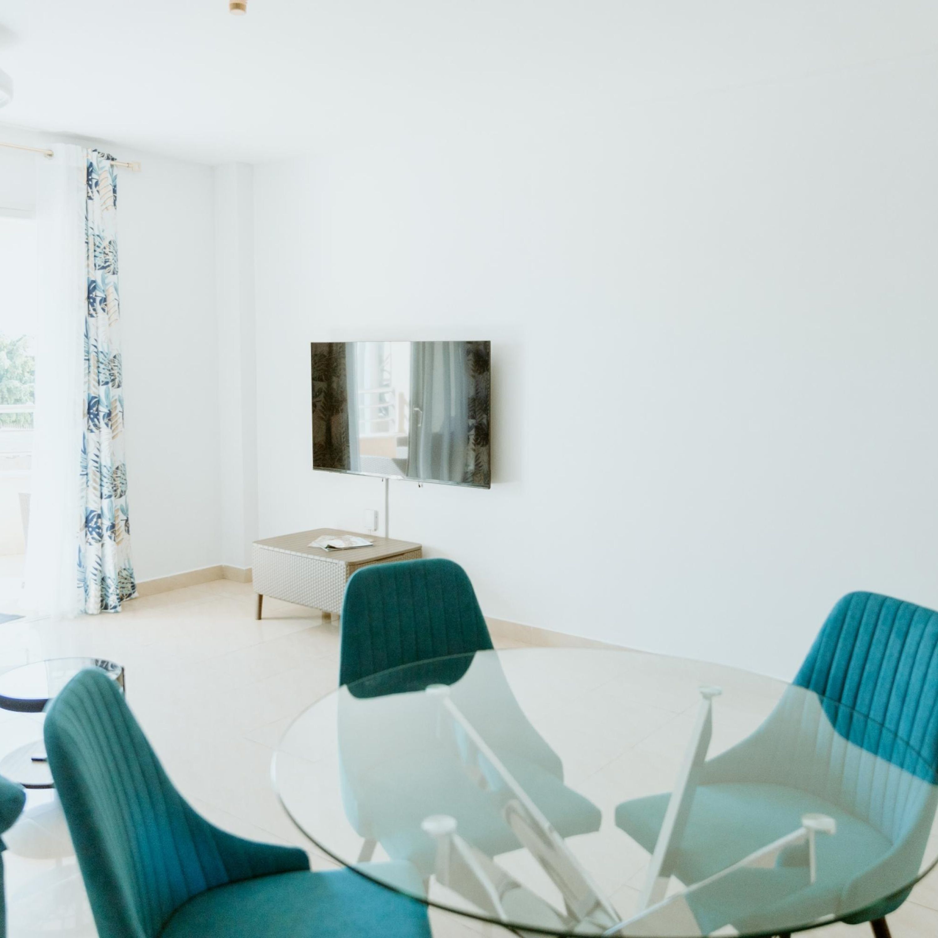 A modern living room with a glass coffee table, teal chairs, a white sofa, a flat-screen TV mounted on the wall, and a window with a curtain.