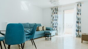 A modern living room with a blue sofa, matching armchairs, a small round table, and a view through a glass door to an outdoor area. - Seaview Splendor 2 Bedroom Modern Comfort (Adeje)