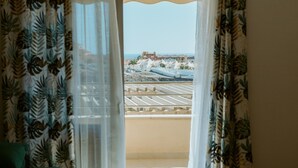 A room with a balcony, white curtains with a tropical leaf pattern, and a view of a coastal area with buildings and a clear sky. - Seaview Splendor 2 Bedroom Modern Comfort (Adeje)