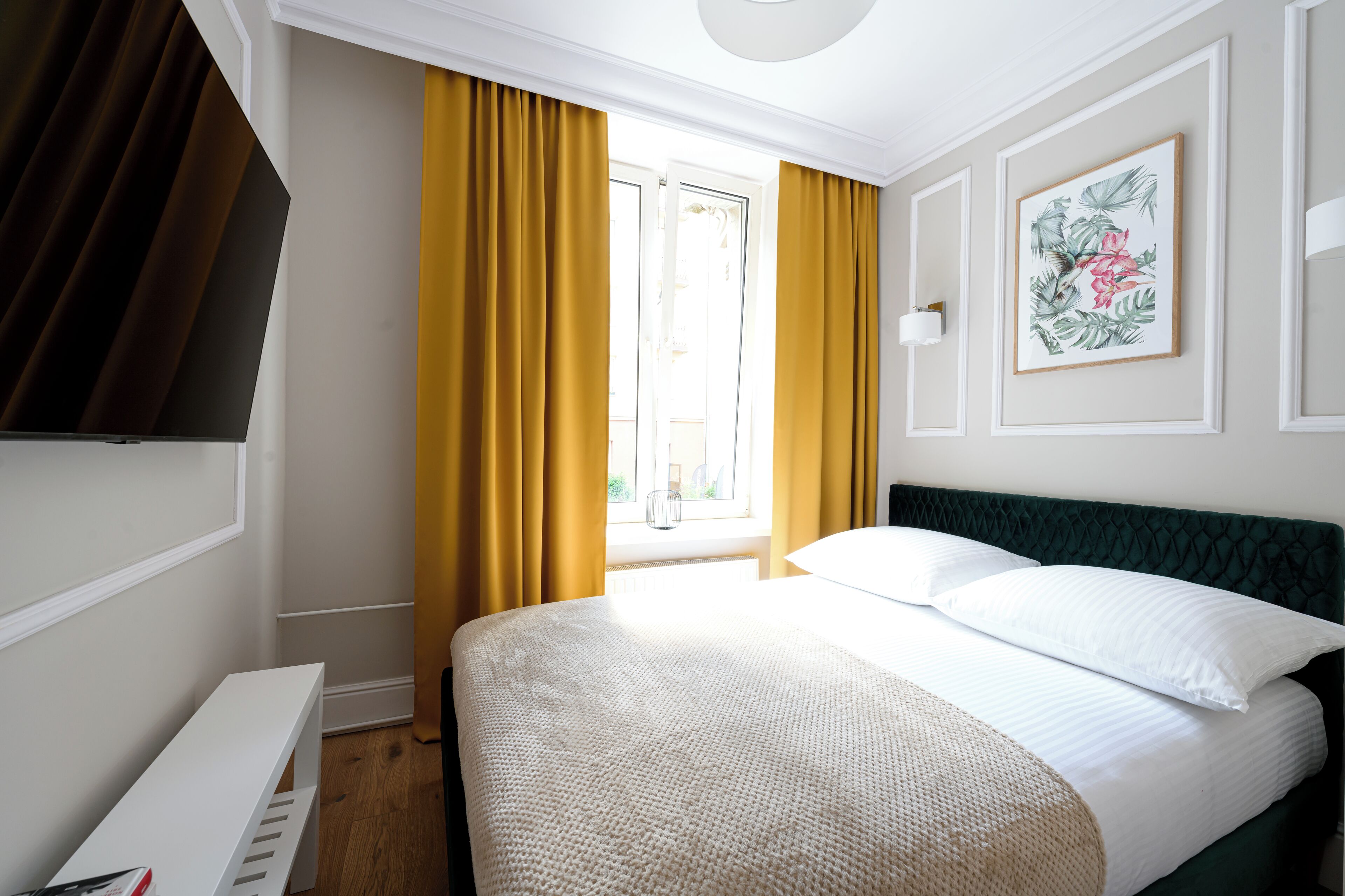 A hotel room with a bed, a desk, a window with yellow curtains, and a framed picture on the wall.