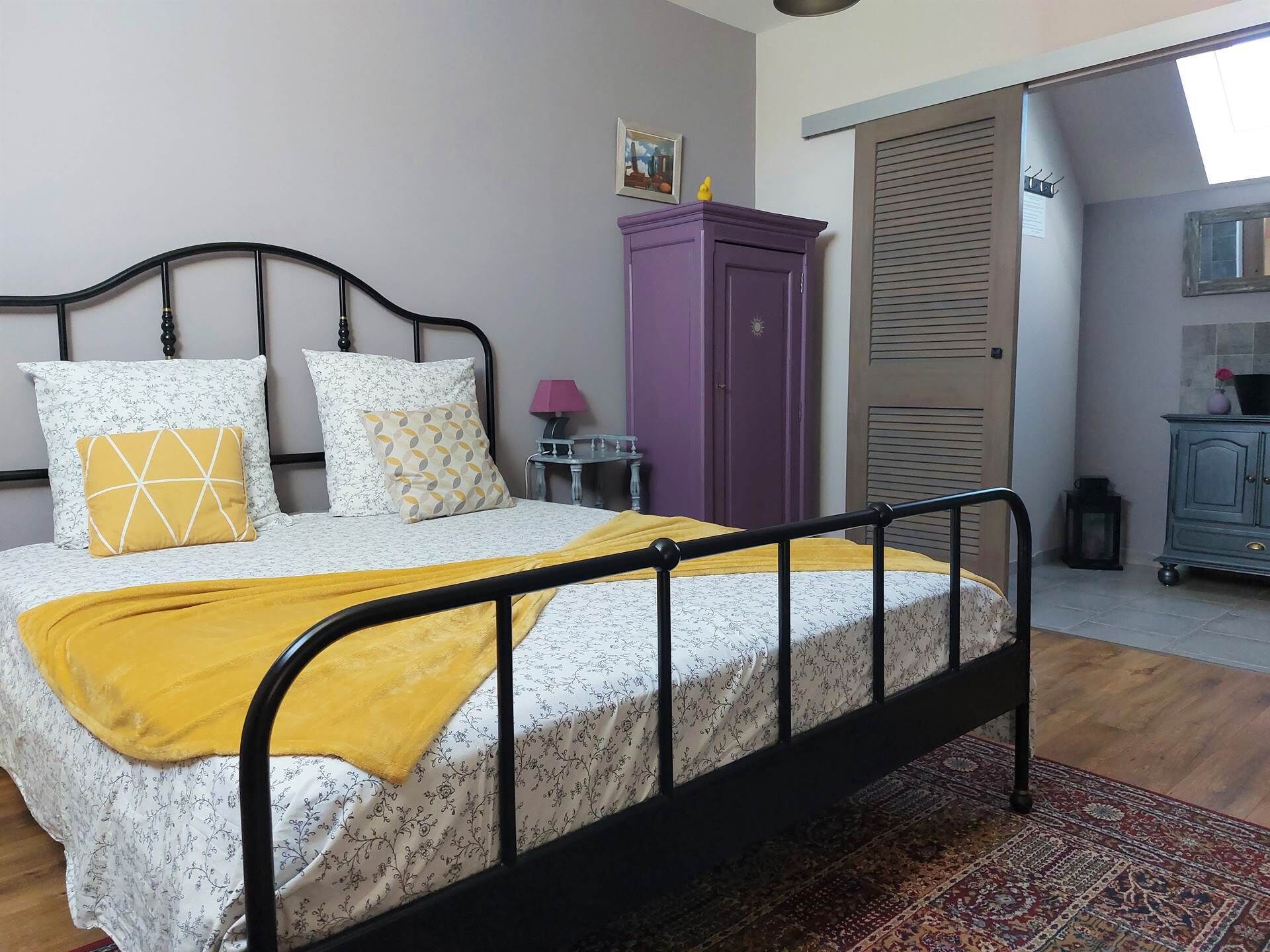 Ein Bett mit schwarzem Metallgestell, weißer Blumenwäsche und einer gelben Tagesdecke. Im Hintergrund steht ein violetter Kleiderschrank und ein kleiner Tisch mit einer Lampe.