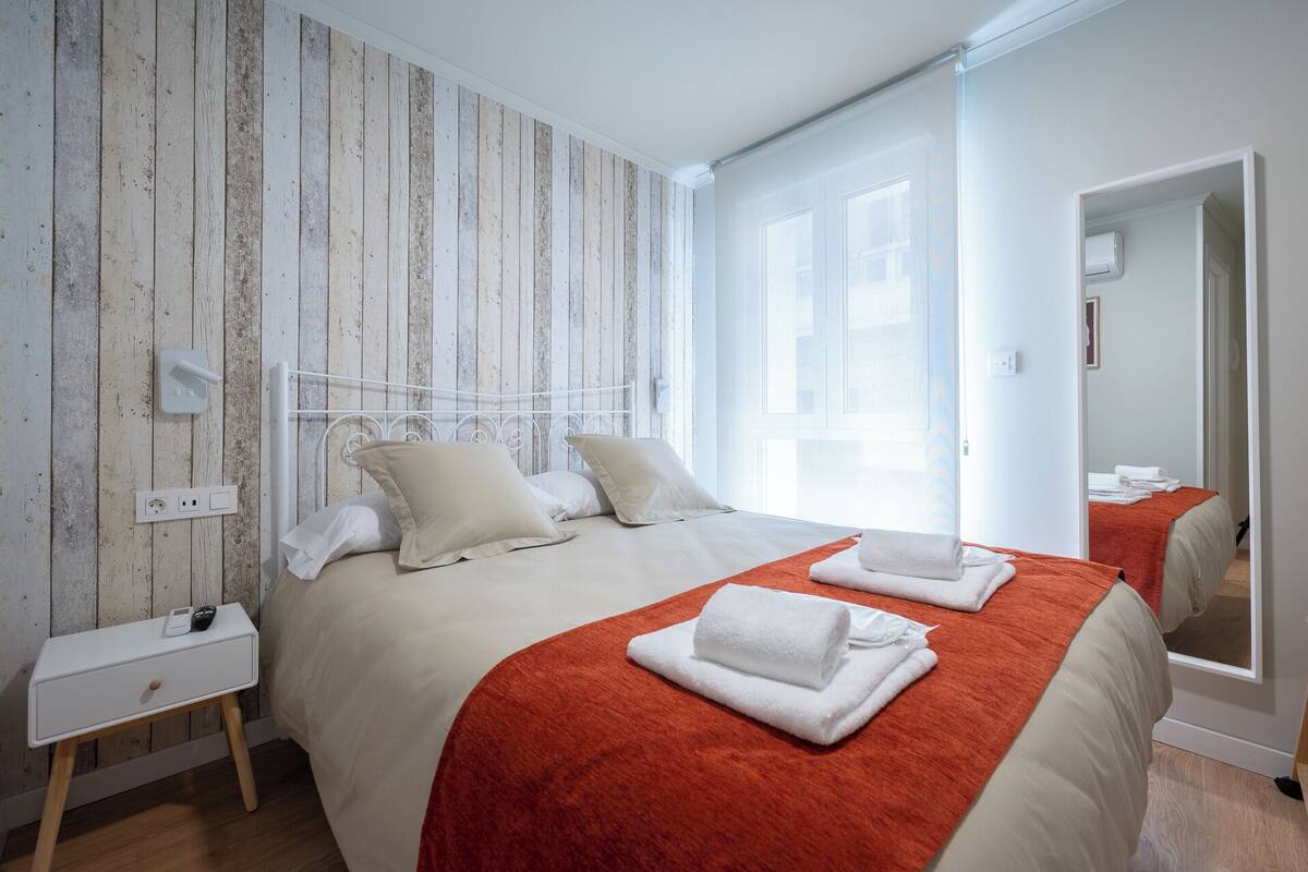 Una habitación de hotel con cama, mesita de noche, enchufe y ventana.