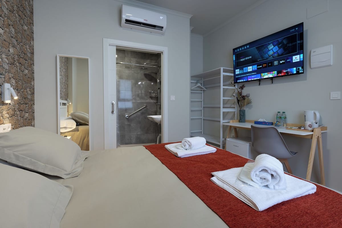 Habitación de hotel con cama, alfombra roja, televisor montado en la pared mostrando varios canales, armario y pequeño escritorio con silla y hervidor.
