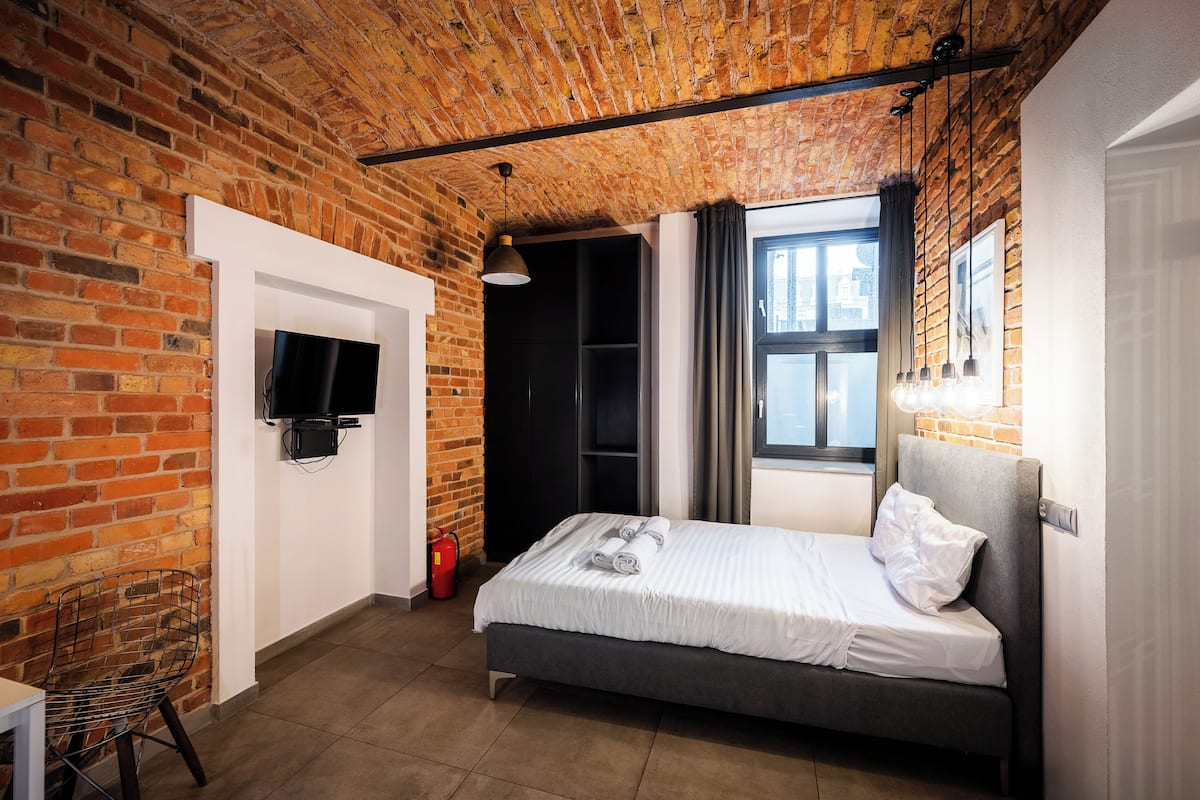 Una habitación con pared de ladrillo, una cama, un televisor, una silla y una ventana con cortinas.