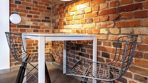 A small, white table with metal wire chairs in a corner with a brick wall and a round light fixture.