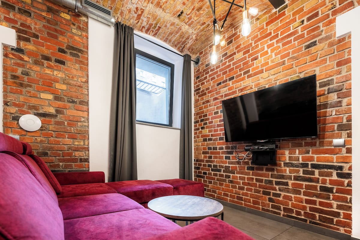 Una sala de estar moderna con una pared de ladrillo, un televisor de pantalla plana grande, un sofá rojo y una pequeña mesa redonda.