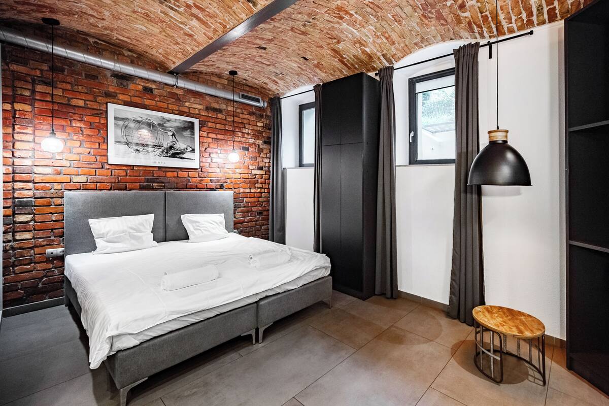 Un dormitorio moderno con pared de ladrillo, una cama, un taburete y una lámpara colgante.