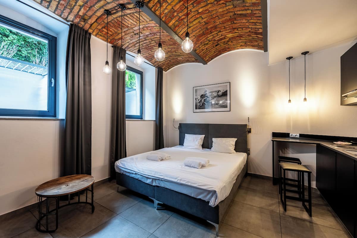 Una habitación de hotel moderna con techo de ladrillo, una cama grande, un escritorio y un ventanal con cortinas.