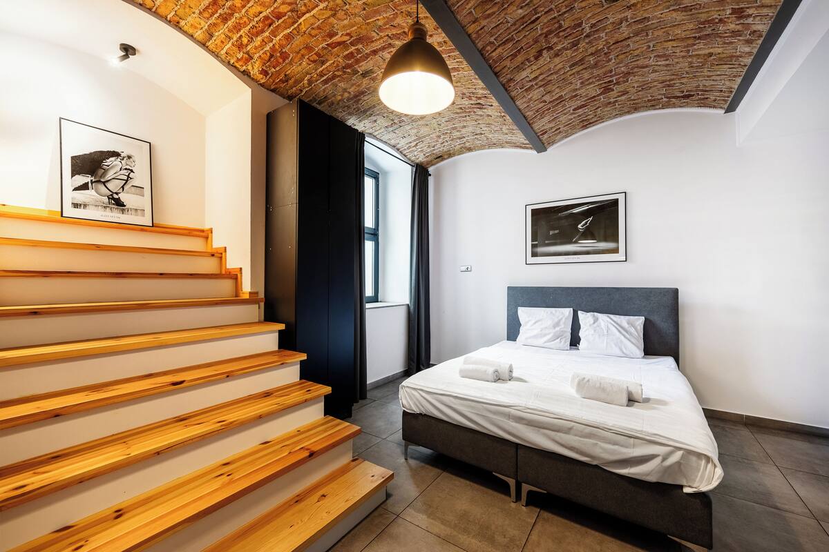 Un dormitorio moderno con una escalera de madera, una cama con ropa de cama blanca y un techo de ladrillo.