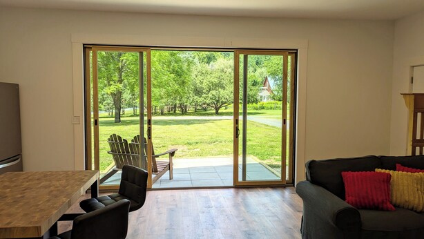 A living room with a sliding glass door leading to a backyard. There is a dining table with chairs, a sofa with cushions, and a television on the left. - Romantic Getaway near Whiteface (Upper Jay)
