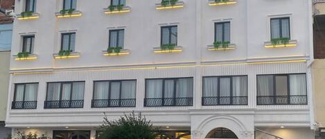 A multi-story hotel with a white facade, balconies with black railings, and green plants in window boxes.